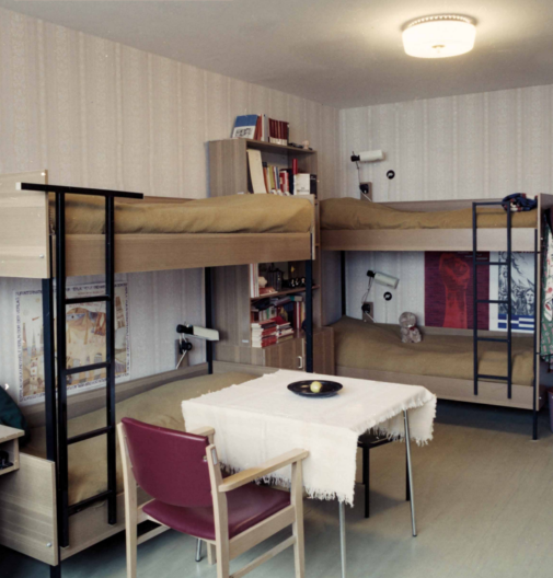 Vierbettzimmer in einem Wohnheim, zwei Doppelstockbetten an den Wänden, in der Mitte des Zimmers ein Tisch mit Stuhl